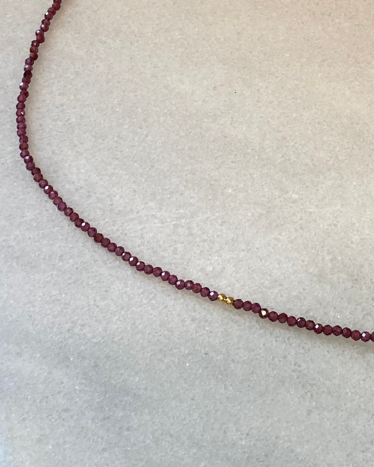 A beaded necklace made of small dark red beads with tiny gold spacer accents and a gold clasp, displayed on a light marble surface.