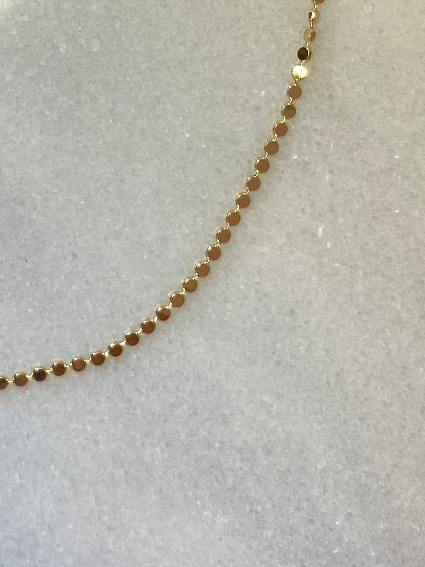 A delicate gold chain necklace with small, faceted beads and an adjustable lobster clasp with an extension chain, displayed on a textured light gray surface.