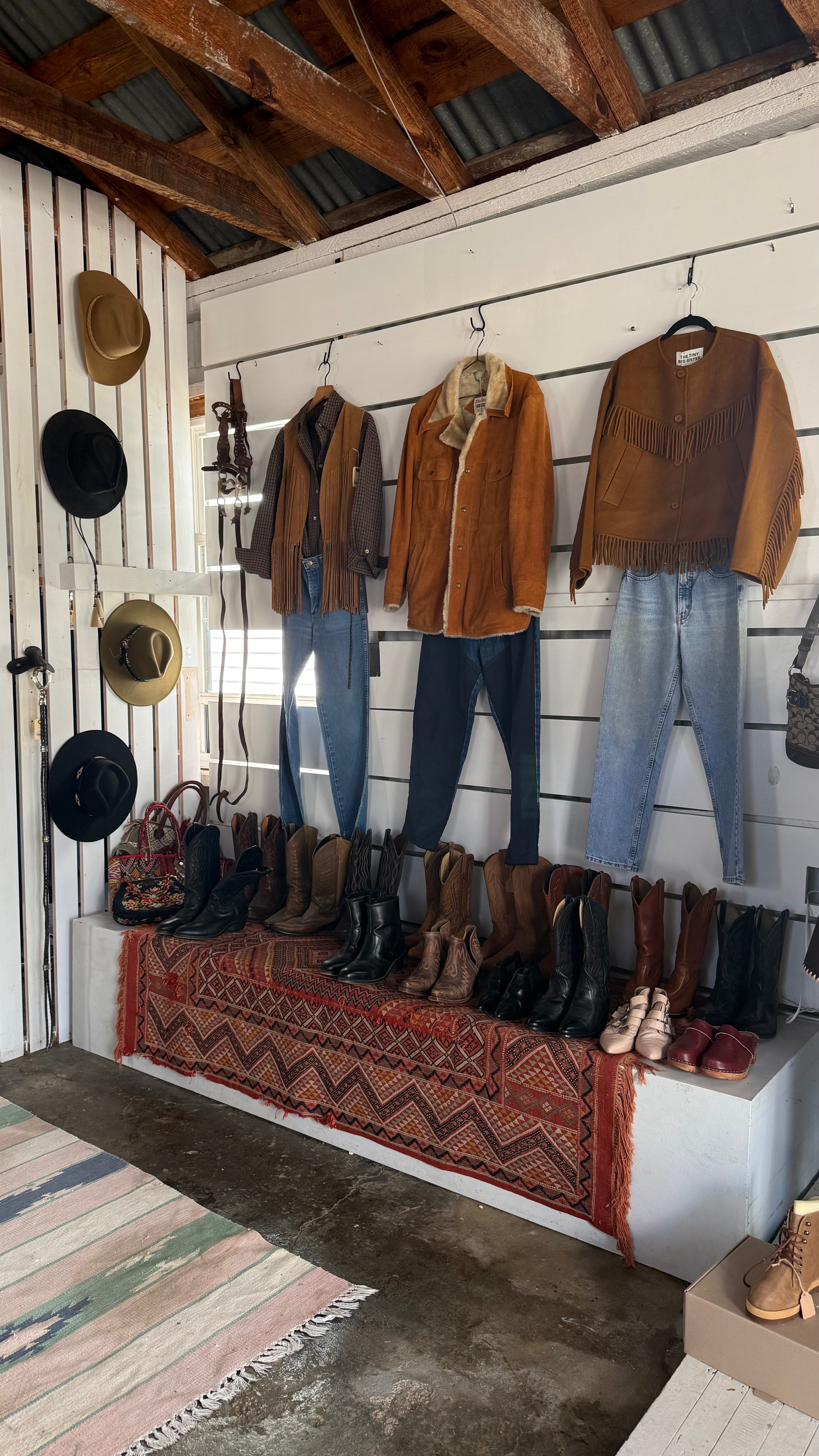 A rustic clothing and boot display inside a barn-style shop. Several brown leather jackets and shirts are hanging on hooks against a white slat wall, paired with jeans and boots.