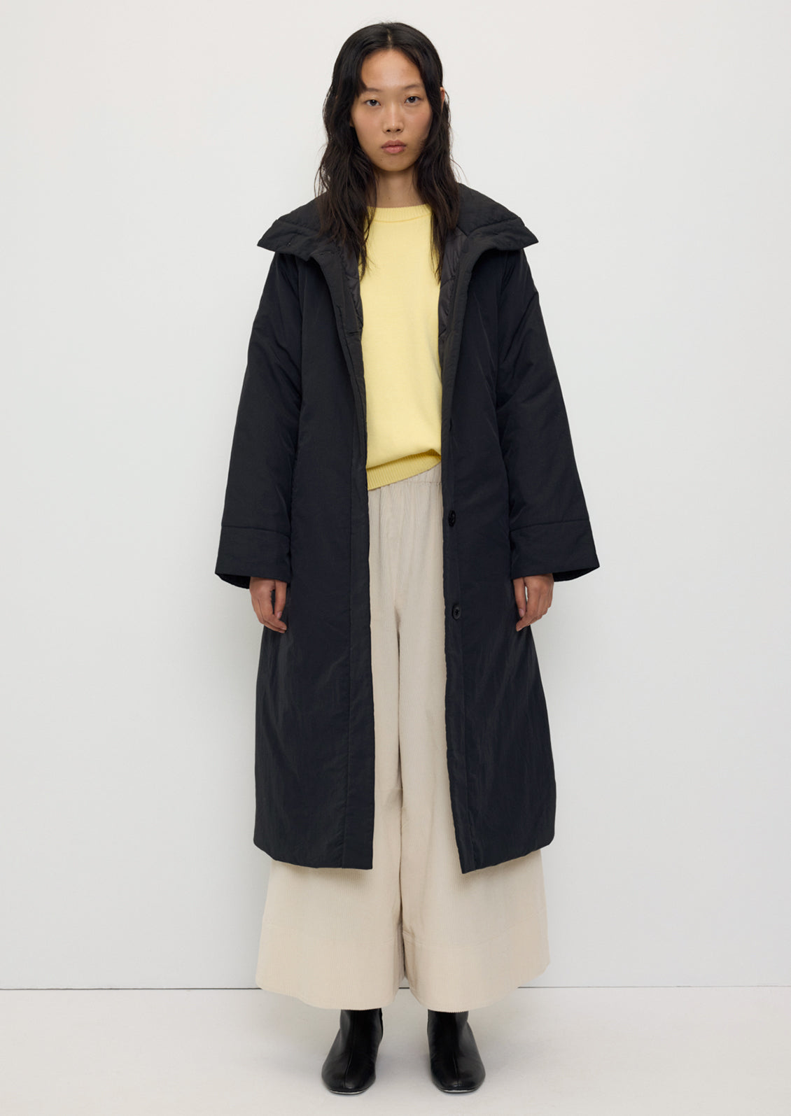 Tender Jacket in Black by Rita Row — worn open over a yellow knit and cream trousers, showing the coat's length and collar