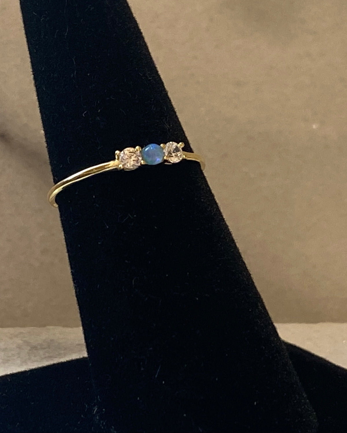 A delicate yellow gold ring with a central round blue gemstone and small clear accent stones, displayed on a black ring stand against a neutral beige background.